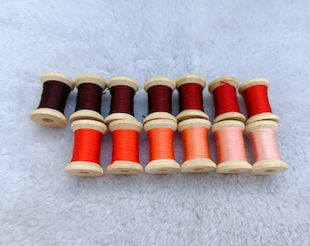 May include: Thirteen wooden spools of thread arranged in two rows, featuring a color gradient that transitions from deep dark brown and maroon on the left to bright red, orange, and soft pastel pink shades on the right side of the collection.