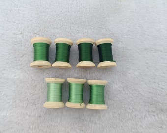May include: Seven wooden spools of thread in varying shades of green. The spools are arranged in two rows, with the top row containing darker green thread and the bottom row featuring lighter green thread. The spools are made of light-colored wood.