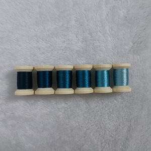 May include: Seven spools of thread in various shades of blue, from dark to light, arranged in a row on a white fuzzy surface.