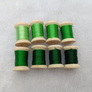 Puede incluir: Ocho carretes de hilo de bordar verde, dispuestos en dos filas de cuatro. Los carretes son de madera y tienen un color marrón claro. El hilo es un degradado de tonos verdes, desde verde claro hasta verde oscuro.
