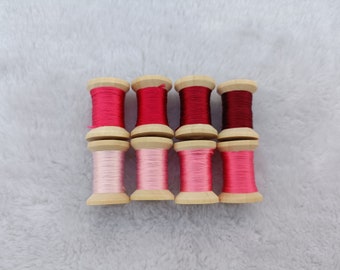 Könnte beinhalten: Acht Garnrollen in verschiedenen Rosétönen und Rottönen. Die Rollen sind aus Holz und haben eine natürliche Oberfläche.