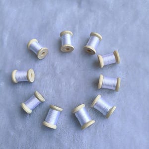 Può includere: Una collezione di dieci piccoli rocchetti di legno disposti in cerchio. Ogni rocchetto è riempito di filo lilla chiaro e ha le estremità in legno naturale. Lo sfondo è un tessuto blu chiaro e morbido.