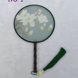 Op de afbeelding: Een ronde, blauw-witte handwaaier met een zwarte steel en een groene kwast. De waaier heeft een ontwerp met een witte magnolia bloem. De tekst "NO 1" is zichtbaar in de linkerbovenhoek van de afbeelding.
