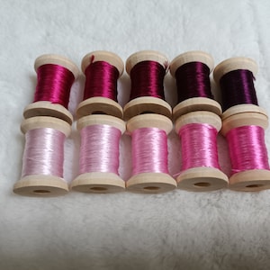 May include: Ten spools of thread in shades of pink and purple. The spools are made of wood and have a natural finish. The thread is wound around the spools and is ready to be used for sewing or embroidery.
