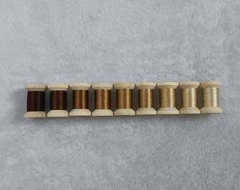 May include: Ten spools of thread in various shades of brown and beige, arranged in a row on a white fuzzy surface.
