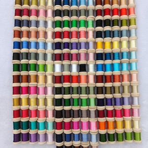 Pode incluir: Uma coleção de 100 carretéis de fio de bordar em uma variedade de cores, dispostos em um padrão de grade. Os carretéis são feitos de madeira e têm uma tampa de metal.