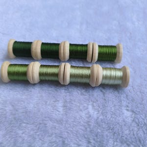 May include: Two wooden spools of thread arranged horizontally. The top spool features a dark forest green thread, while the bottom spool displays a lighter, sage green thread. Both spools have a natural light wood finish with circular ends.