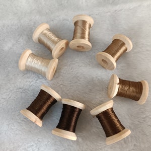 May include: Eight spools of thread, four brown and four beige, are arranged in a circle on a white fuzzy surface. The spools are made of wood and have a simple design.