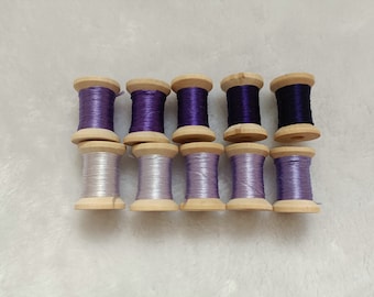 May include: Ten spools of thread in shades of purple and lavender, wound on wooden bobbins.