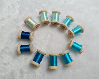 May include: Ten wooden spools of embroidery thread arranged in a circular pattern. The thread colors range from pale mint green to various shades of bright cyan, sky blue, and deep royal blue, all featuring a smooth, shiny, and reflective finish.