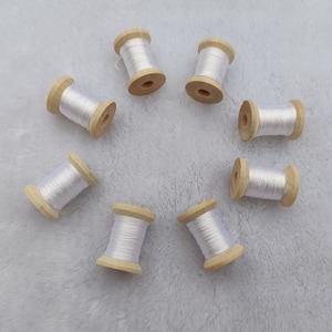 Puede incluir: Ocho carretes de hilo blanco enrollados alrededor de bobinas de madera. Los carretes están dispuestos en círculo sobre una superficie blanca y peluda.