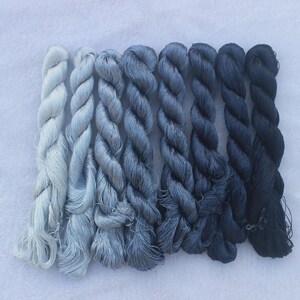 May include: A collection of eight skeins of hand-dyed embroidery floss in shades of blue, ranging from a very light blue to a dark navy blue.