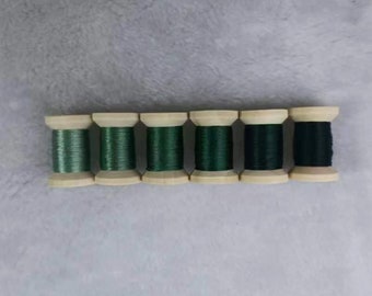 May include: Six wooden spools of thread in varying shades of green, from light to dark, are arranged in a row. The thread is tightly wound around the wooden spools, creating a neat and organized appearance. The spools are set against a soft, gray background.