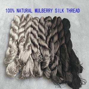 Può includere: Una collezione di 100% NATURAL MULBERRY SILK THREAD disposta in una sfumatura di colori dal beige chiaro e grigio argento al grigio antracite scuro e nero. I fili sono intrecciati in matasse individuali, mostrando una consistenza liscia e lucente.