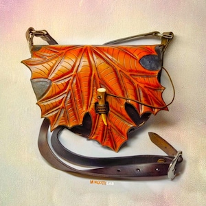 Puede incluir: Un bolso de hombro de cuero marrón con un gran diseño de hoja de arce. El bolso tiene una correa con hebilla y un pequeño acento de hueso.