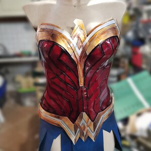 Op de afbeelding: Een rood, goud en zilveren Wonder Woman kostuum-bovenstuk met een blauwe rok. Het bovenstuk heeft een gedetailleerd ontwerp met een grote, gouden en zilveren ster in het midden.