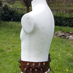 Puede incluir: Un cinturón de cuero marrón con detalles metálicos y pequeñas bolsas adjuntas. El cinturón se lleva en un maniquí blanco.