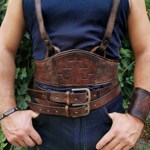 May include: A brown leather belt with a decorative buckle and a matching leather strap with a Celtic knot design. The belt is worn over a pair of blue jeans and a black tank top.