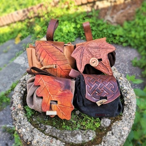 May include: Four leather pouches with leaf and Celtic knot designs. The pouches are in shades of brown and tan, with details like wooden toggles and leather straps. The pouches are displayed in a stone planter with greenery.