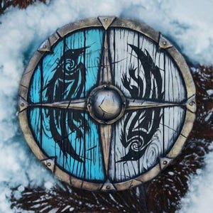 May include: A round wooden shield with a metal rim. The shield is painted in half blue and half white. Each half features a black stylized bird design.