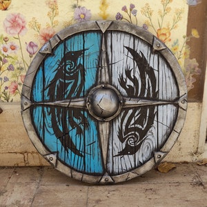 May include: A round wooden shield with a metal rim. The shield is painted in half blue and half white with black stylized bird designs on each side.