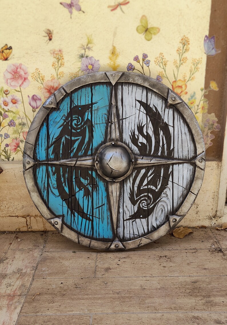 May include: A round wooden shield with a metal rim. The shield is painted in half blue and half white with black stylized bird designs on each side.