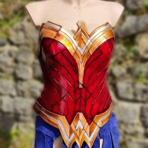 Op de afbeelding: Een rood en gouden Wonder Woman kostuum-bovenstuk met een gedetailleerd ontwerp. Het bovenstuk is gemaakt van een glanzend materiaal en heeft een gesculpteerde look.