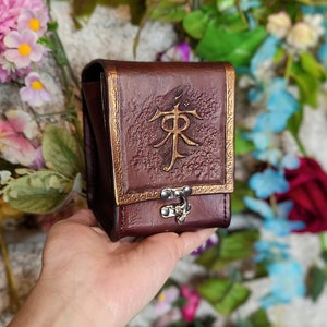 May include: A brown leather dice bag with a gold embossed design and a metal clasp. The bag is decorated with a stylized symbol.