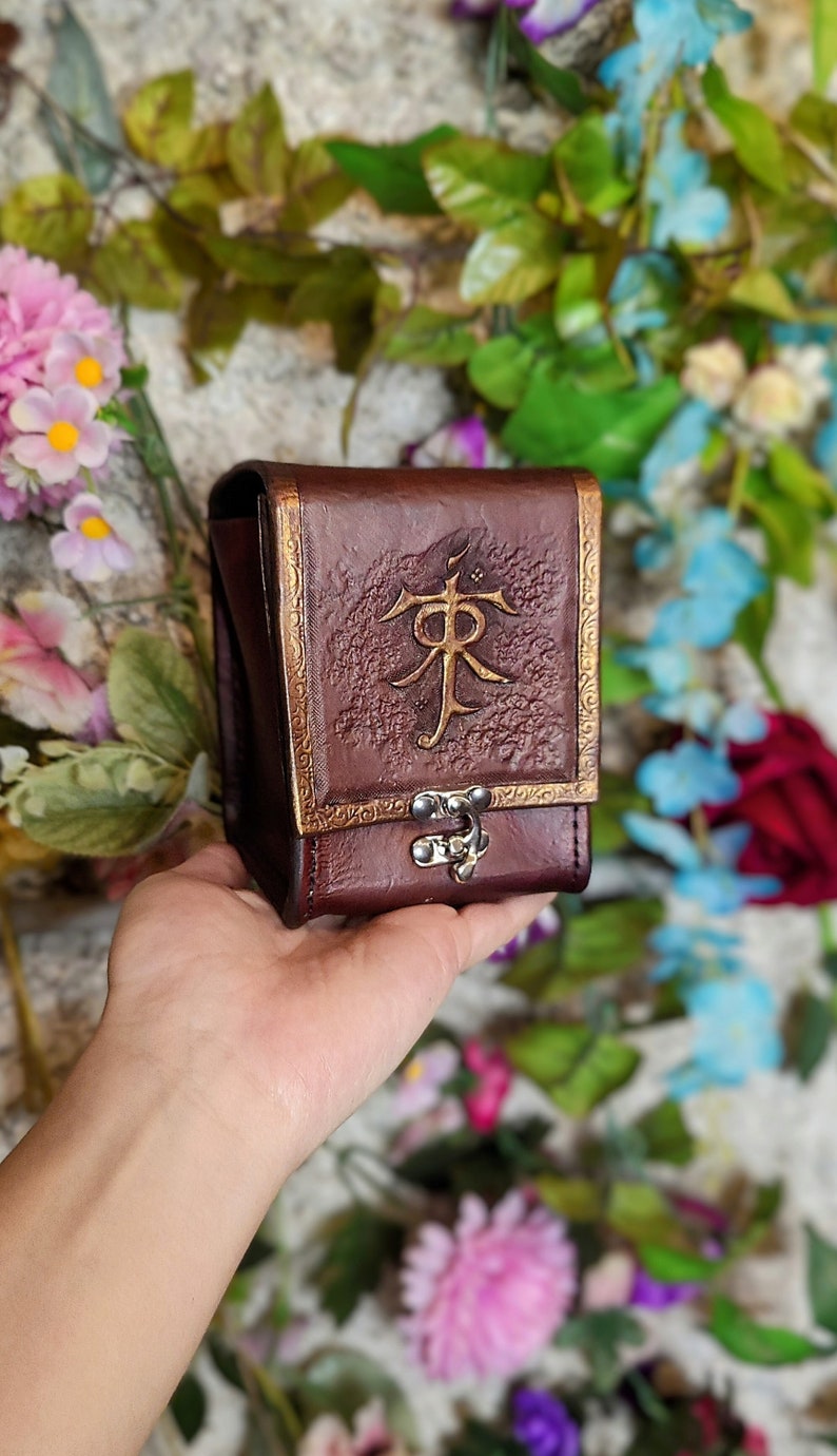May include: A brown leather dice bag with a gold embossed design and a metal clasp. The bag is decorated with a stylized symbol.