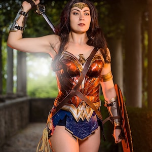 Op de afbeelding: Een vrouw verkleed als Wonder Woman, draagt een rood, goud en bruin kostuum met een blauwe rok, en houdt een zwaard en schild vast. Ze staat in een bosachtige omgeving.
