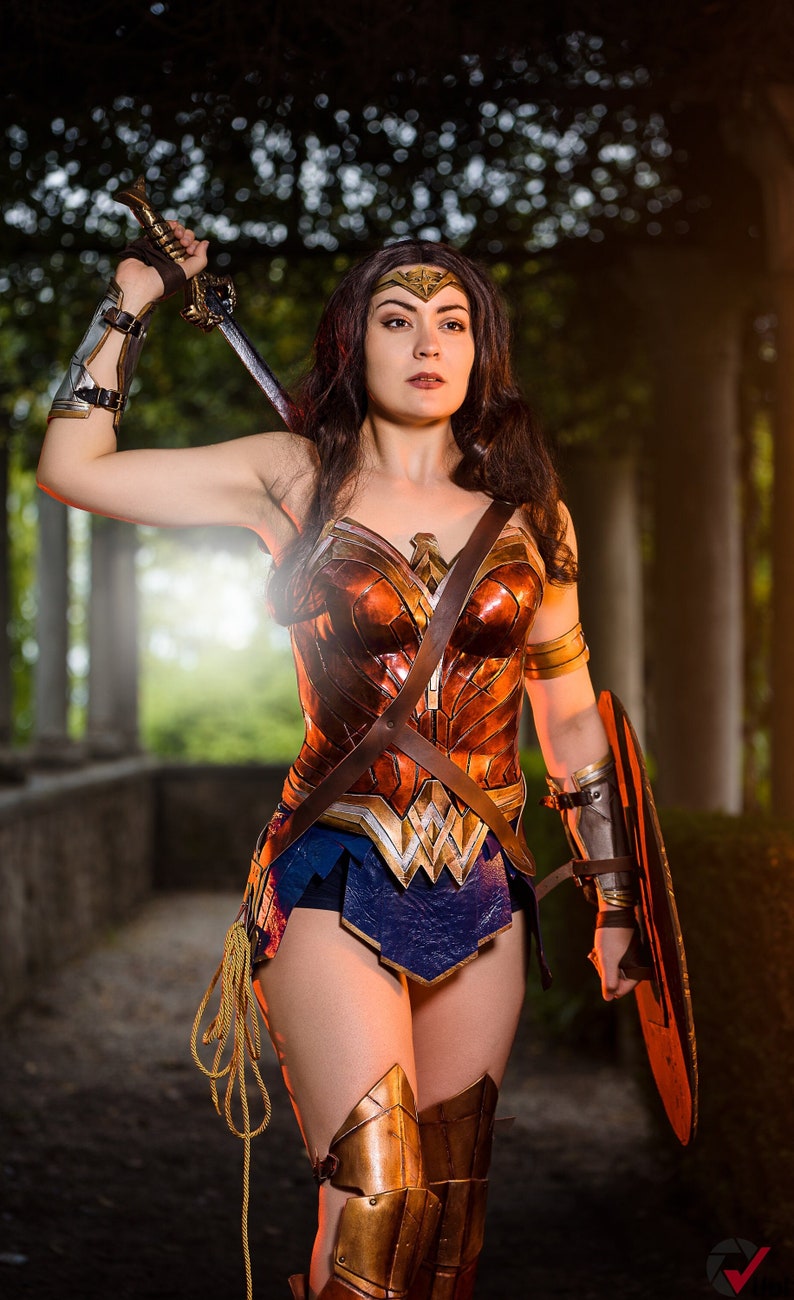 Op de afbeelding: Een vrouw verkleed als Wonder Woman, draagt een rood, goud en bruin kostuum met een blauwe rok, en houdt een zwaard en schild vast. Ze staat in een bosachtige omgeving.
