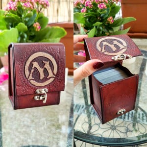 May include: A brown leather card deck box with a silver clasp and a gold embossed "M" logo. The box is open and shows a deck of cards inside.