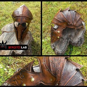 May include: A brown leather helmet with a dragon scale design. The helmet has a face mask with a pointed nose and eye holes. The helmet is decorated with brass rivets and studs. The helmet is sitting on a wooden stump.