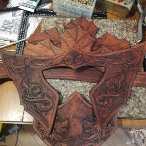 May include: Close-up of a leather craft project, featuring multiple pieces of brown leather with intricate floral designs carved into the surface. The leather pieces are arranged on a wooden surface.