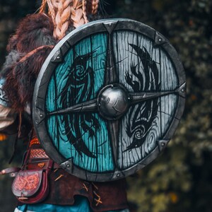 May include: A round shield with a black and white design of stylized birds on a blue and white background. The shield is made of wood and has a metal rim.