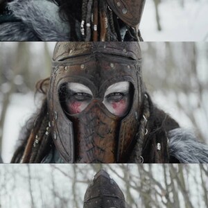 May include: A person wearing a brown leather helmet with intricate carvings and a face mask. The helmet has a pointed top and is decorated with metal studs. The person has long, dark hair and is wearing a fur-lined coat.