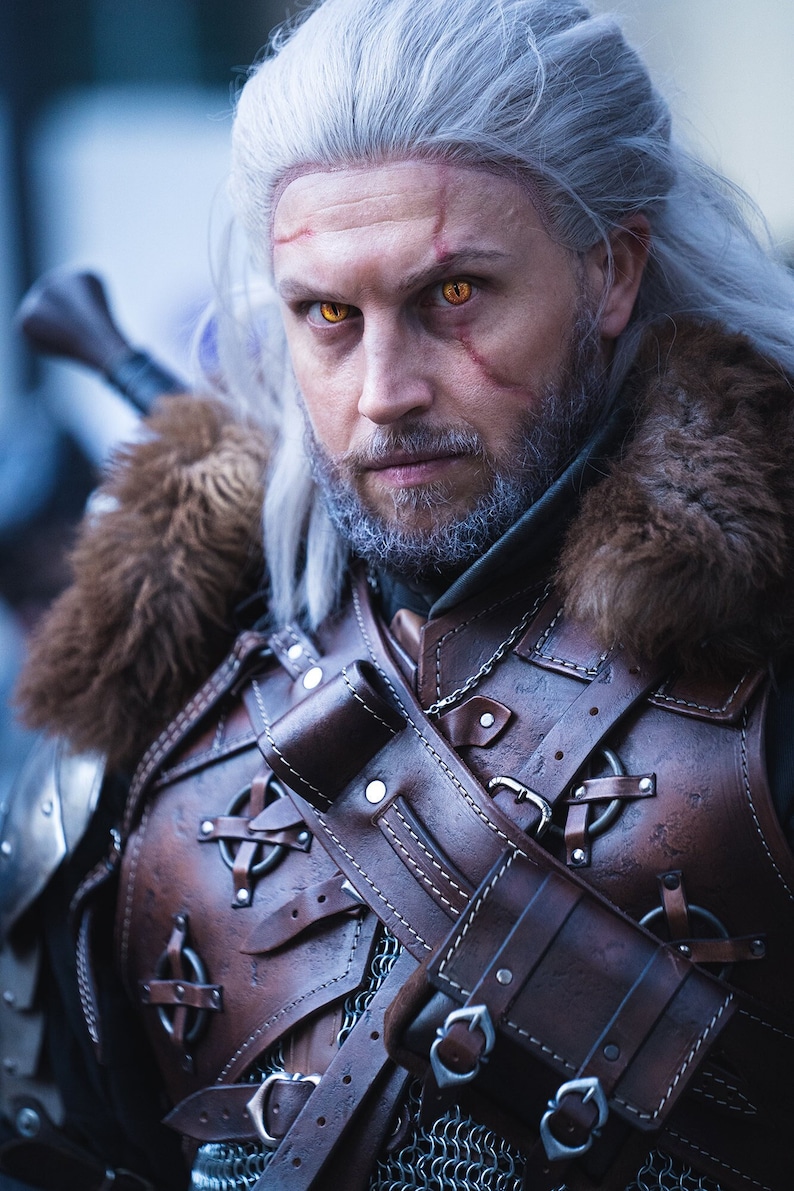 May include: A man dressed as Geralt of Rivia from the Witcher video game series. He is wearing a brown leather armor with a fur collar, a white wig, and has yellow contact lenses.