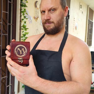 May include: A man wearing a black apron holds a small, brown leather box with a silver clasp. The box has a design of two stylized animals on the front.
