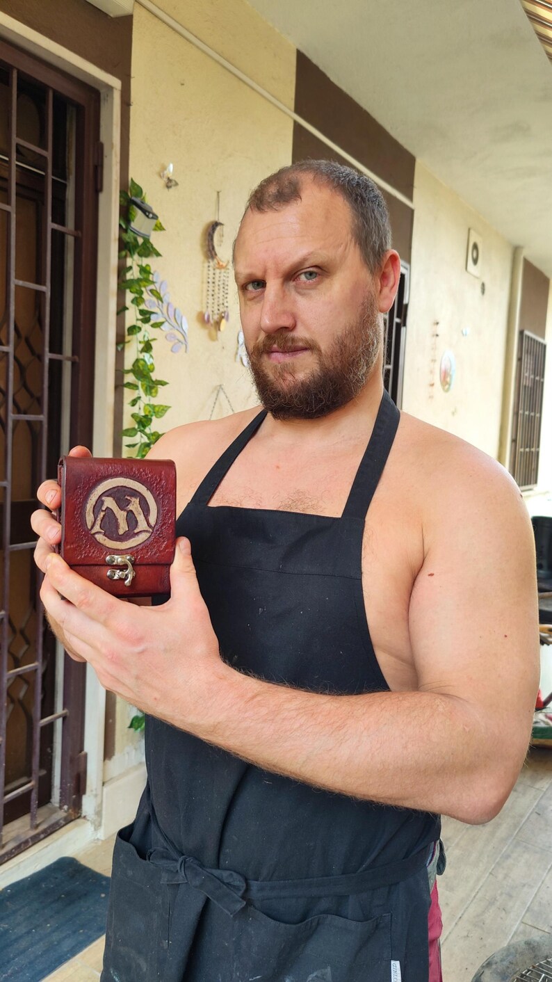 May include: A man wearing a black apron holds a small, brown leather box with a silver clasp. The box has a design of two stylized animals on the front.