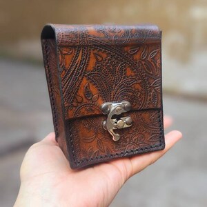 May include: A small, brown leather box with a paisley pattern and a silver clasp. The box is held in a hand.