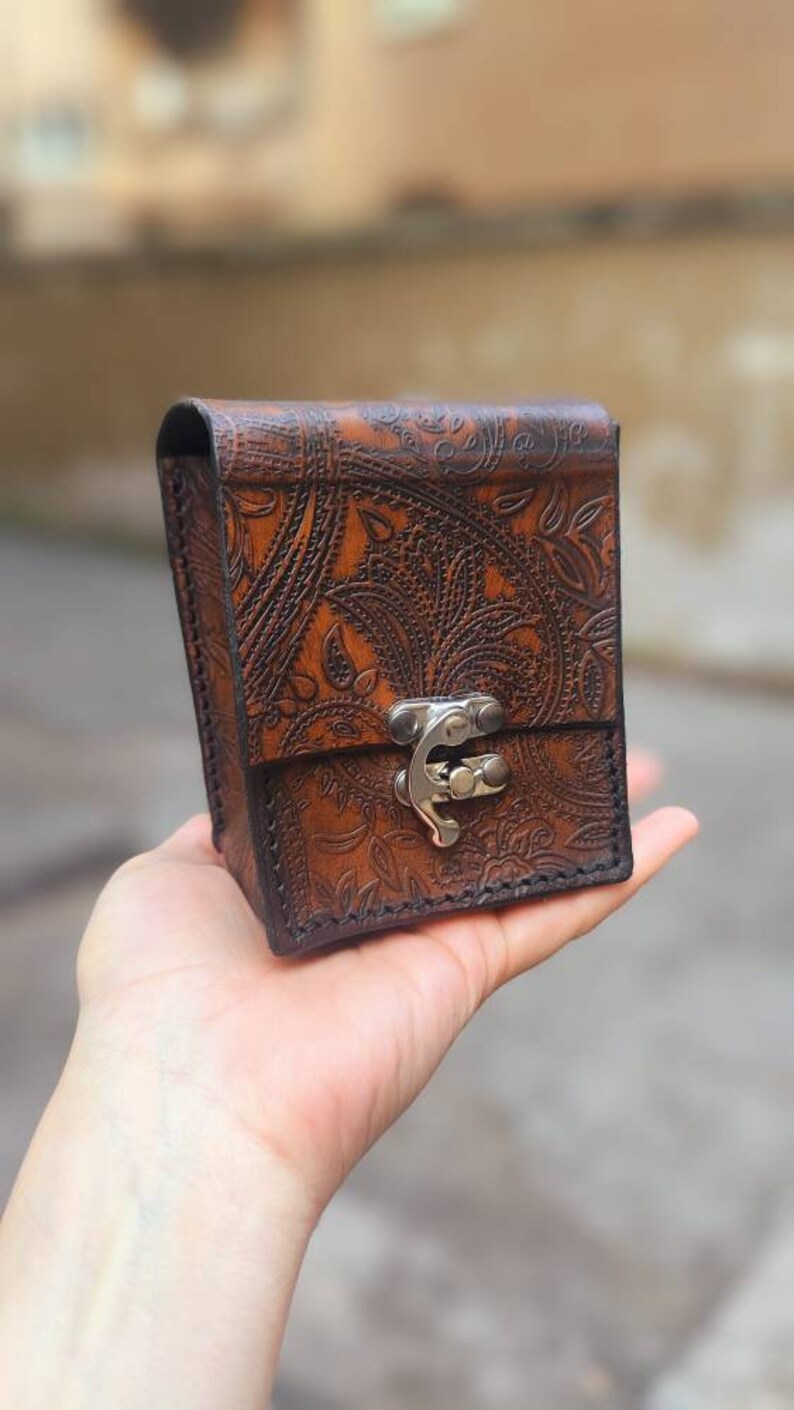 May include: A small, brown leather box with a paisley pattern and a silver clasp. The box is held in a hand.