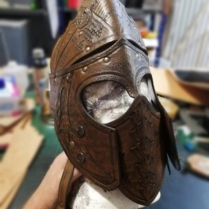 May include: A brown leather helmet with intricate embossed details and metal accents. The helmet has a visor and a strap.