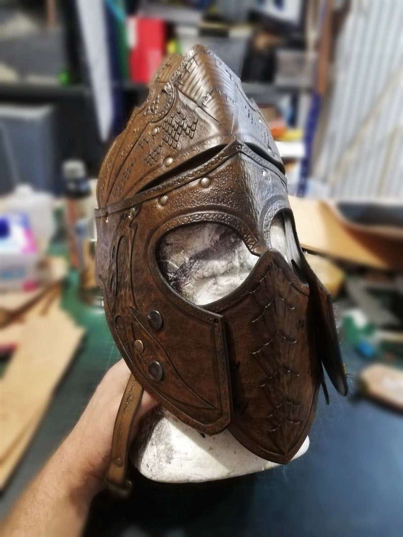 May include: A brown leather helmet with intricate embossed details and metal accents. The helmet has a visor and a strap.