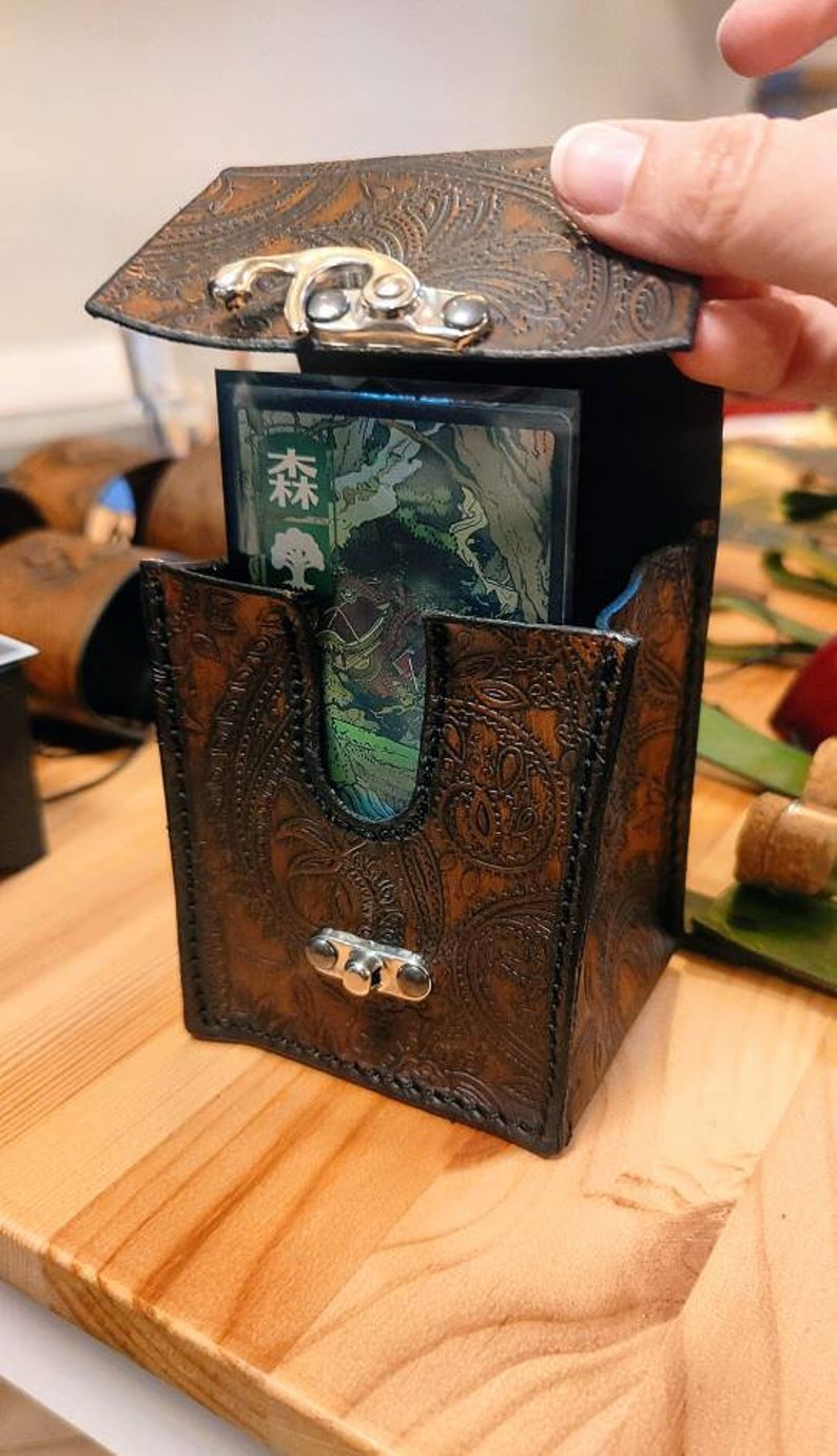 May include: A brown leather card deck box with a paisley pattern and a metal clasp. The box is open and a card with Japanese text is visible inside.