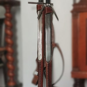 May include: Two swords with brown leather sheaths, hanging on a wall. The sheaths have decorative stitching and the swords have metal accents.