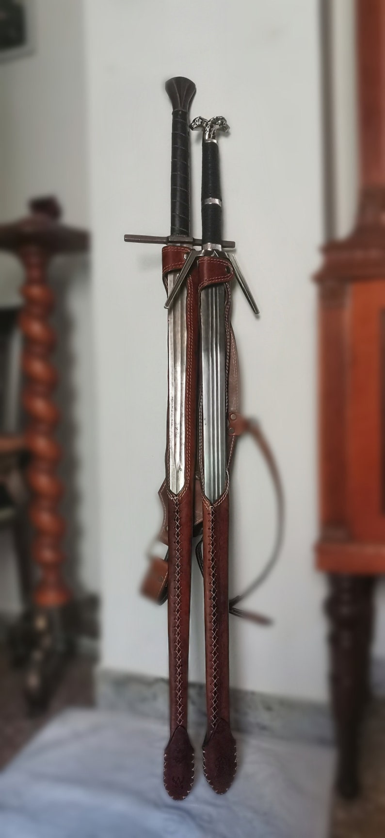 May include: Two swords with brown leather sheaths, hanging on a wall. The sheaths have decorative stitching and the swords have metal accents.