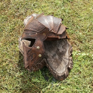 May include: A brown leather helmet with a textured surface and metal accents. The helmet has a pointed top and a face mask with a decorative design.