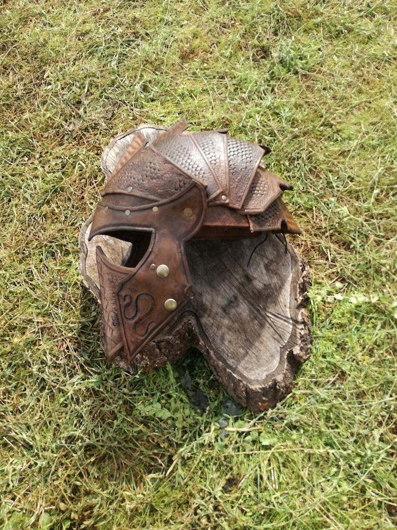 May include: A brown leather helmet with a textured surface and metal accents. The helmet has a pointed top and a face mask with a decorative design.