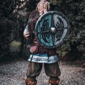 May include: A woman in a warrior costume with a shield on her back. The shield is round and has a blue and white design with a black bird on it. The woman is wearing a brown leather belt with a pouch and a blue and white tunic. She is standing on a gravel path.