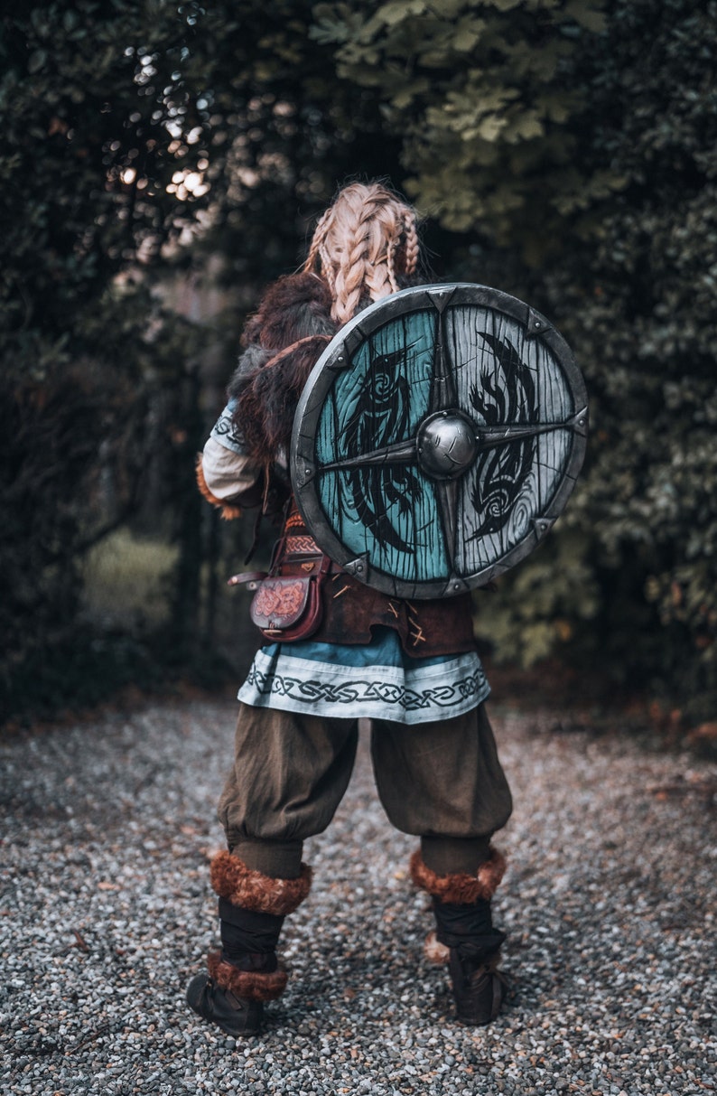 May include: A woman in a warrior costume with a shield on her back. The shield is round and has a blue and white design with a black bird on it. The woman is wearing a brown leather belt with a pouch and a blue and white tunic. She is standing on a gravel path.